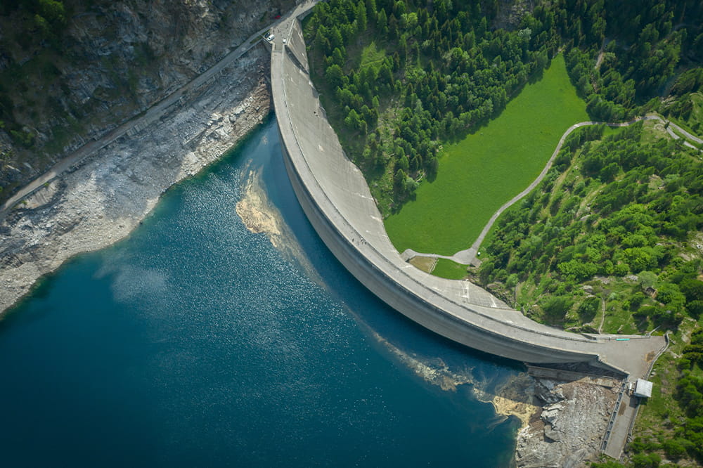 Bird's eye view of a dam dividing the image, with water in the lower left corner of the image and green meadow in the upper right corner.
