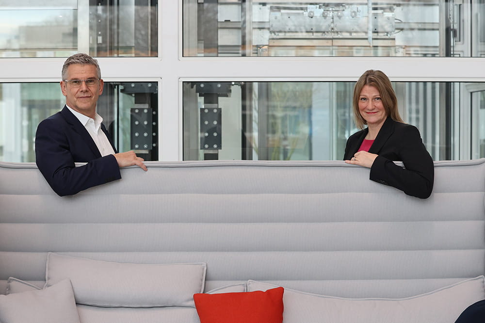 Two people sit on a couch and look back at the camera with a smile.