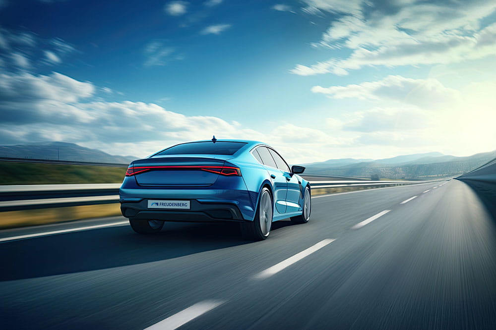 A blue sedan drives on an empty highway under a partly cloudy sky with mountains visible in the distance.