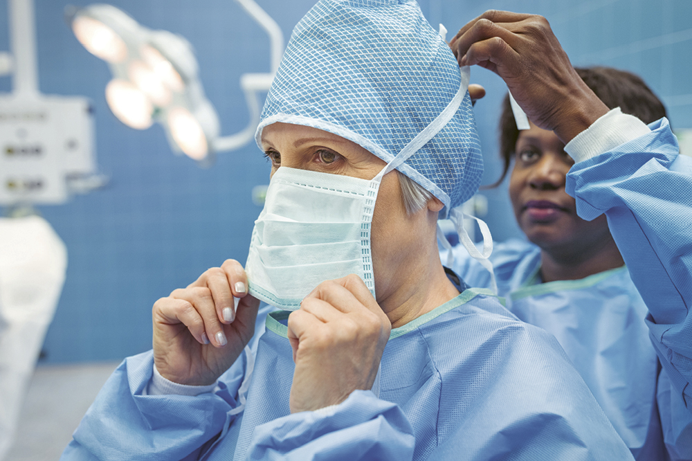 Scene of two doctors in surgical gowns - one ties a face mask to the other's head.