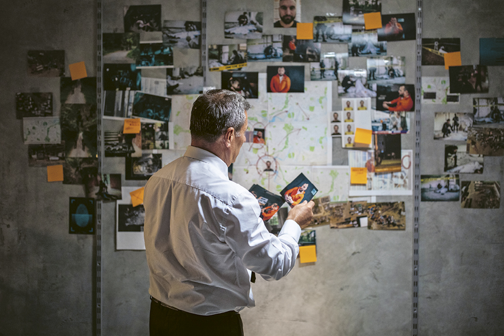 Man in front of a pinboard with pictures about an investigation with inmates and a map of the city