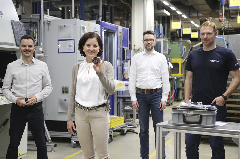 Daniel Uhl, Tanja Heislitz, Christian Kleinke and Christoph Schäfer stand in a production hall, look into the camera and Tanja Heislitz shows a DIAvent.
