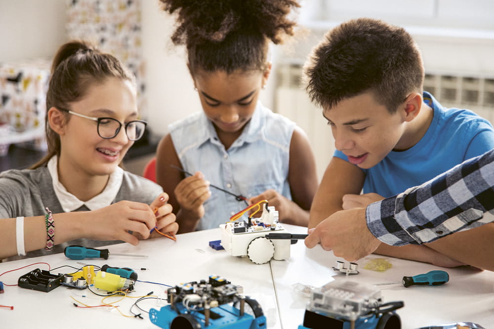 Three children sit around a table and tinker with a small robot. Copyright: iStock: M_a_y_a