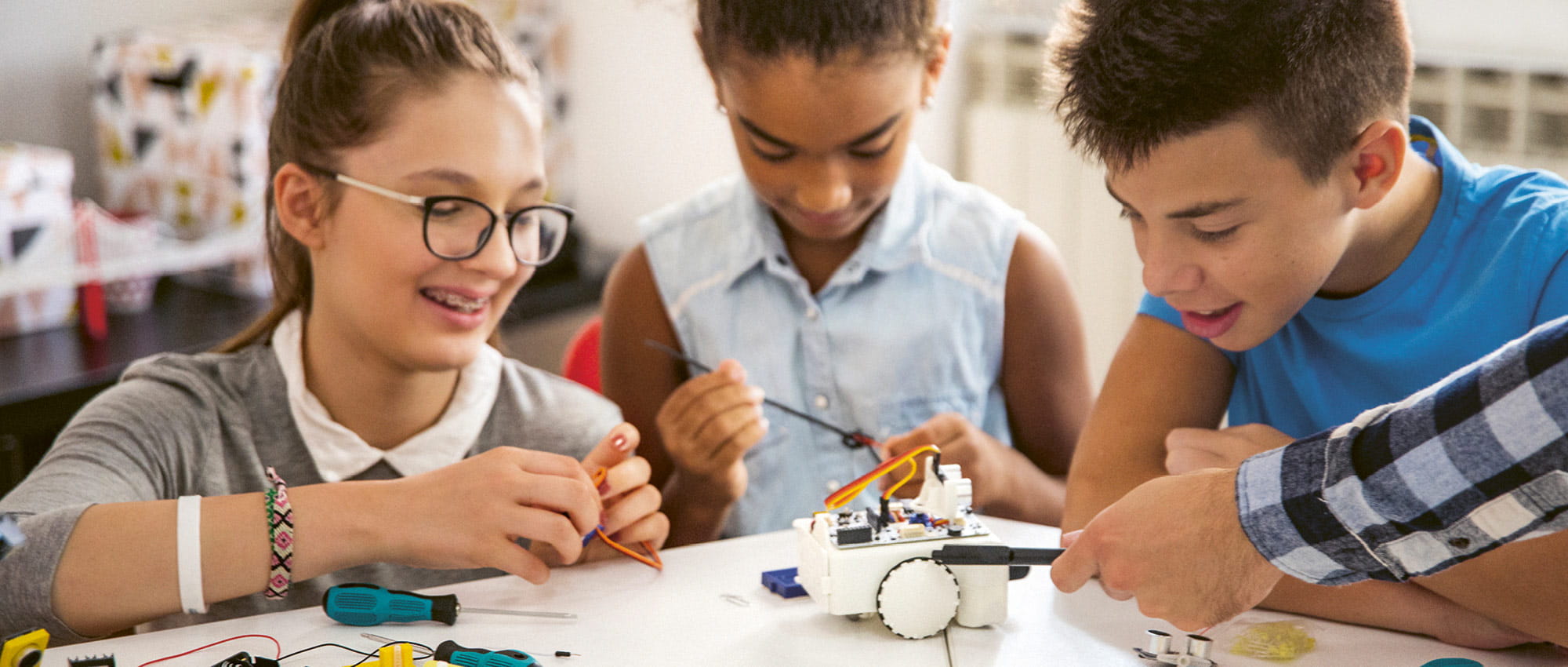 Three children sit around a table and tinker with a small robot. Copyright: iStock: M_a_y_a