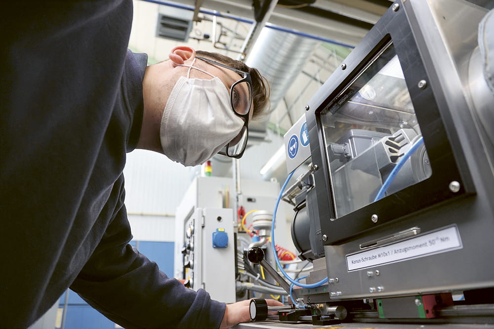 A man in a mask looks at the test bench of a machine in a factory.