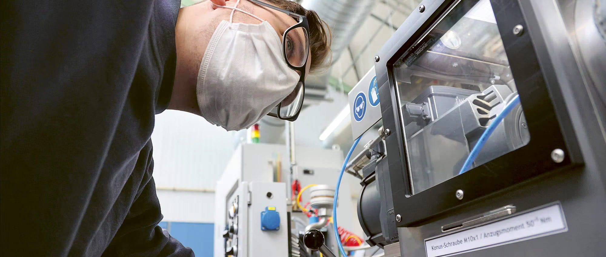 A man in a mask looks at the test bench of a machine in a factory.