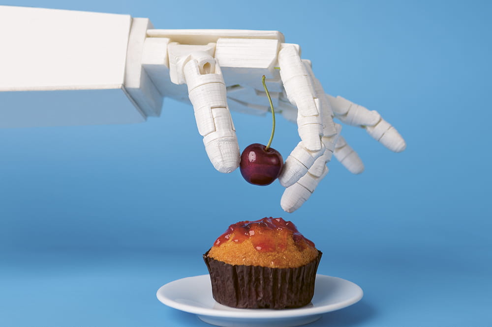 A white robot hand grabs a cherry and places it on a muffin. 