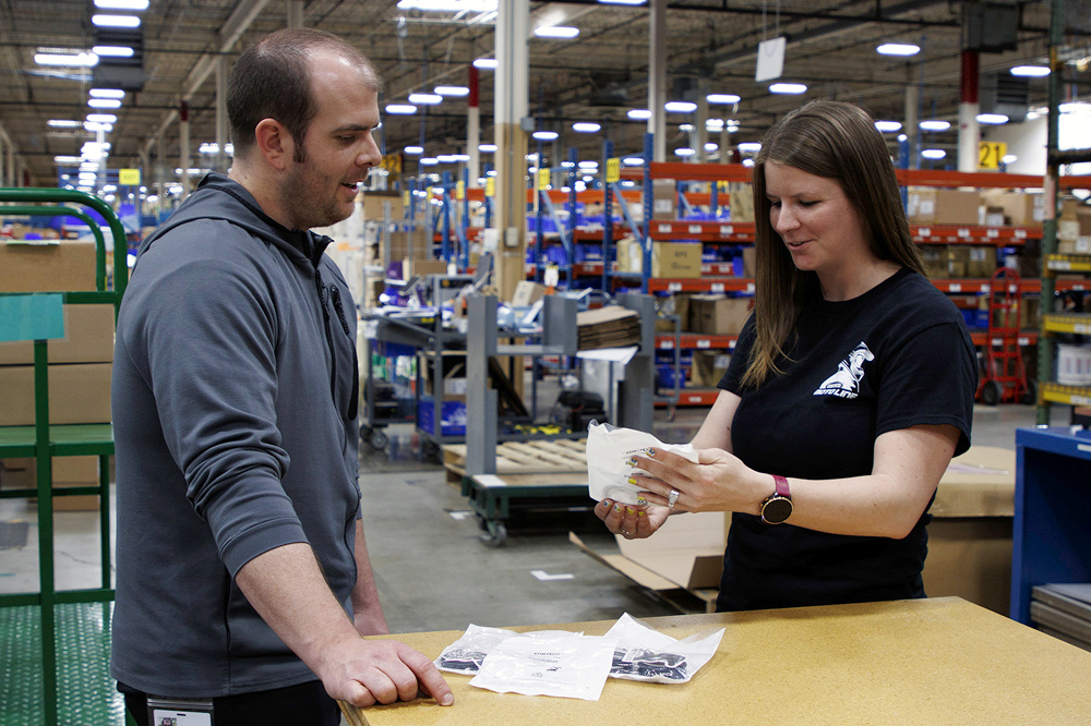A man stands in a warehouse and advises a customer who is holding a fork seal in her hand. 