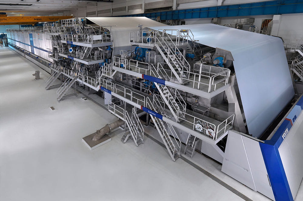 Industrial paper manufacturing machine in a modern factory hall, where a continuous roll of white paper is produced, with metal stairs, safety railings, control platforms, and heavy industrial equipment.