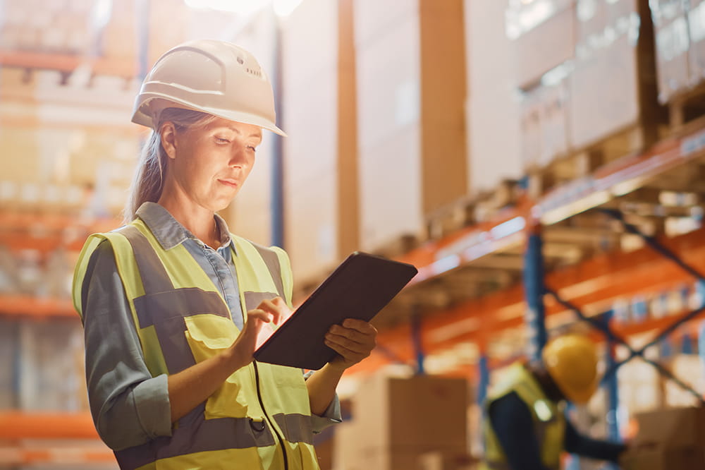 Woman with safety vest and helmet with storage shelfs in background looking at her tablet