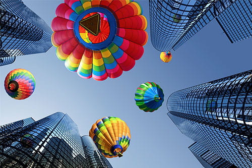 Five colorful hot air balloons rising between glass skyscrapers against blue sky