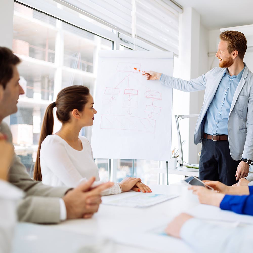 Male in business suit points on a drawing on a flip chart while several people are listening