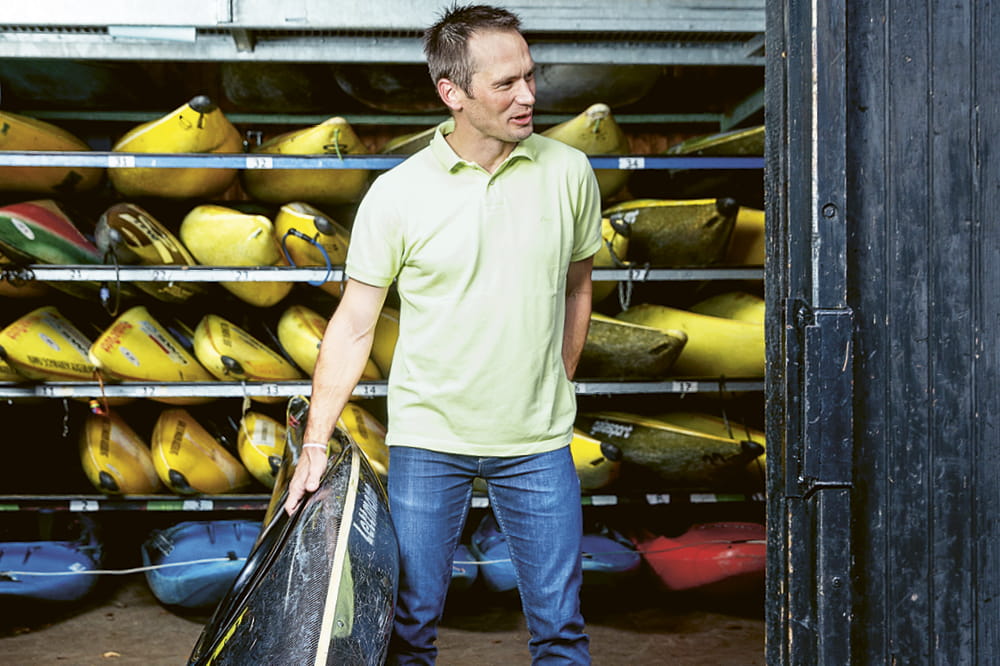 Thomas Schmitt stehend mit einem Kayak vor einem Schuppen voller Kajaks.