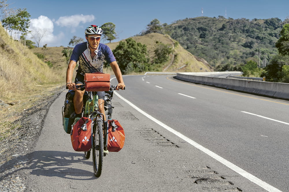 Der Fahrradfahrer Basti fährt auf seinem mit Satteltaschen bepacktem Fahrrad eine Straße entlang