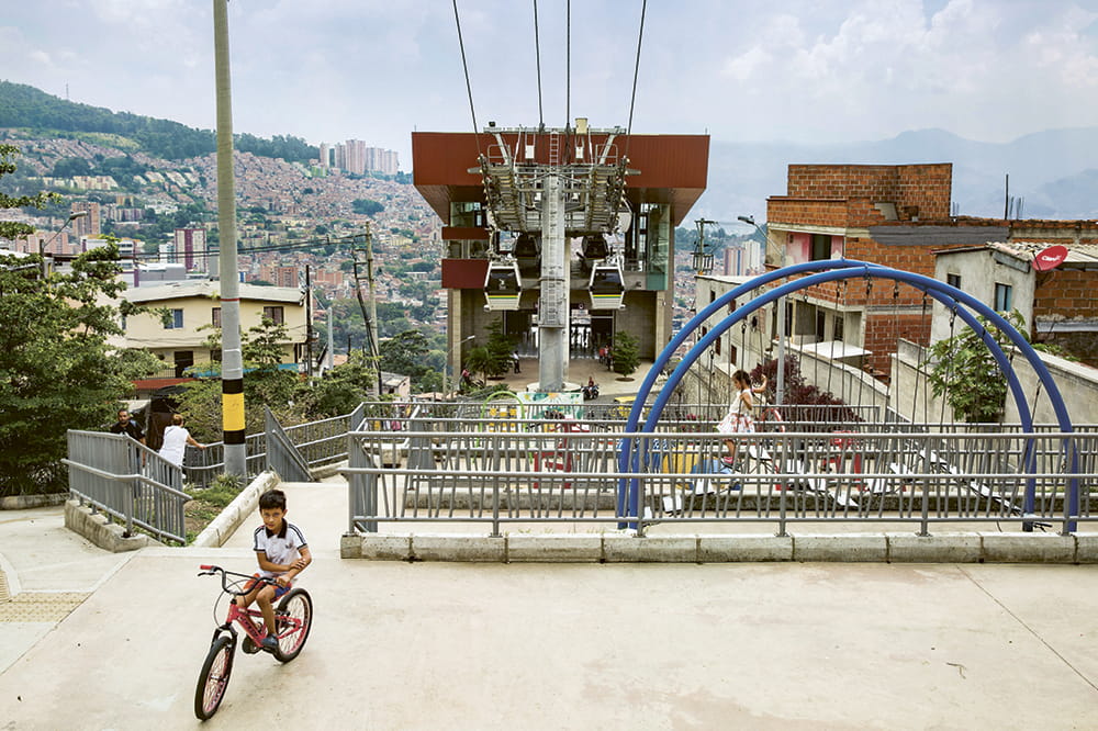 Junge auf einem Fahrrad und spielende Kinder in einem Wohngebiet von Medellin mit einer Seilbahnstation mittendrin.