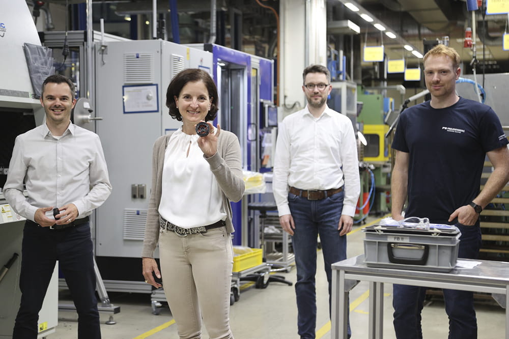 Daniel Uhl, Tanja Heislitz, Christian Kleinke und Christoph Schäfer stehen in einer Produktionshalle, schauen in die Kamera und Tanja Heislitz zeigt ein DIAvent.