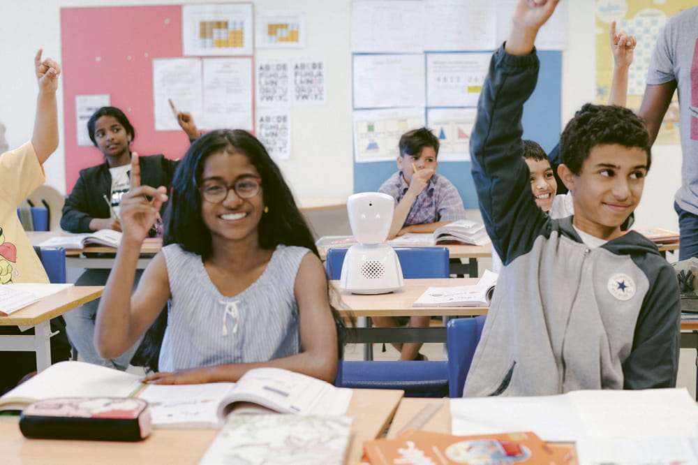 Kinder, die in einem Klassenzimmer sitzen, lächeln und heben die Hände. Zwischen ihnen steht auf einem freien Tisch ein kleiner weißer Roboter.