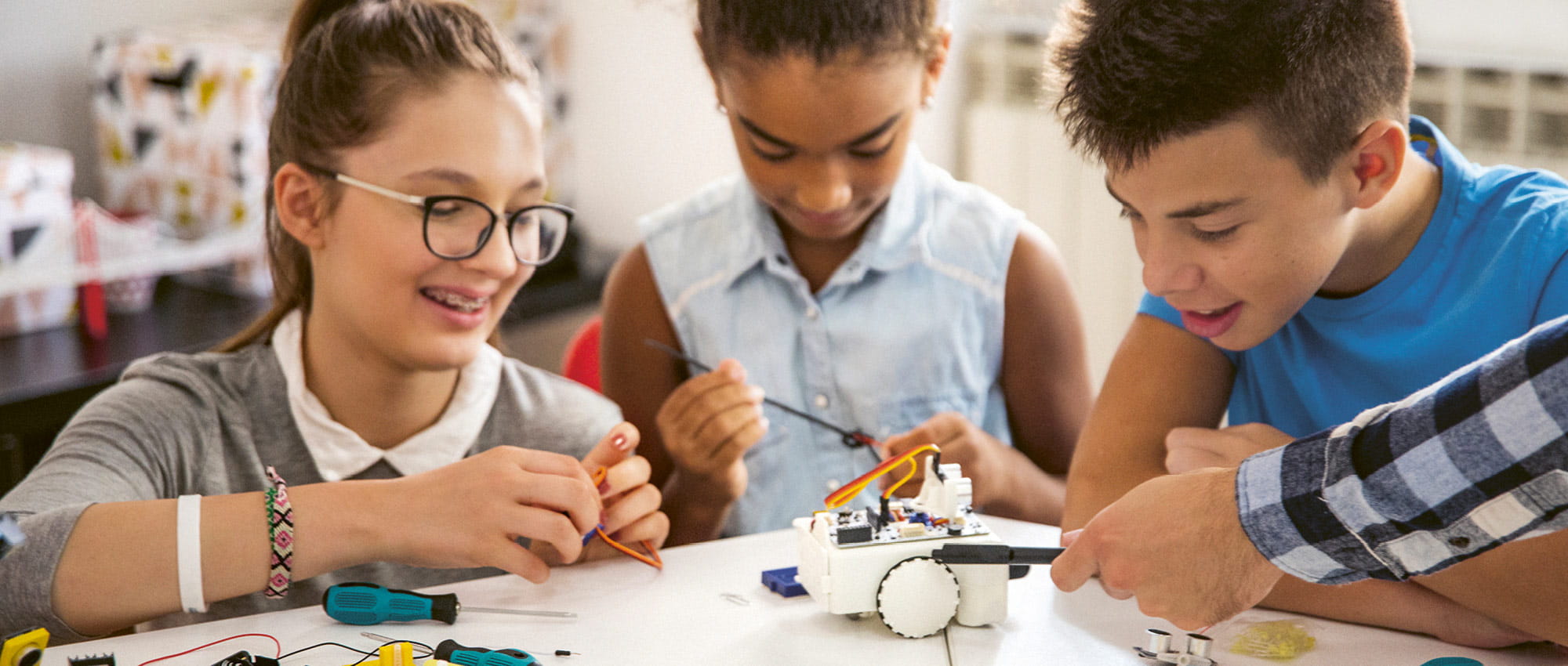 Drei Kinder sitzen um einen Tisch und basteln an einem kleinen Roboter. Copyright: iStock: M_a_y_a