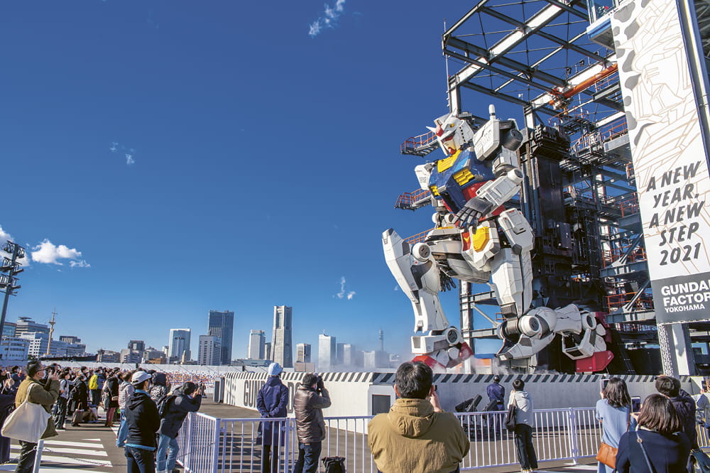 Haushoher Roboter kniet auf Plattform mit Skyline im Hintergrund und Menschenmenge, die ihn fotografiert.  Copyright: Dick Thomas Johnson 