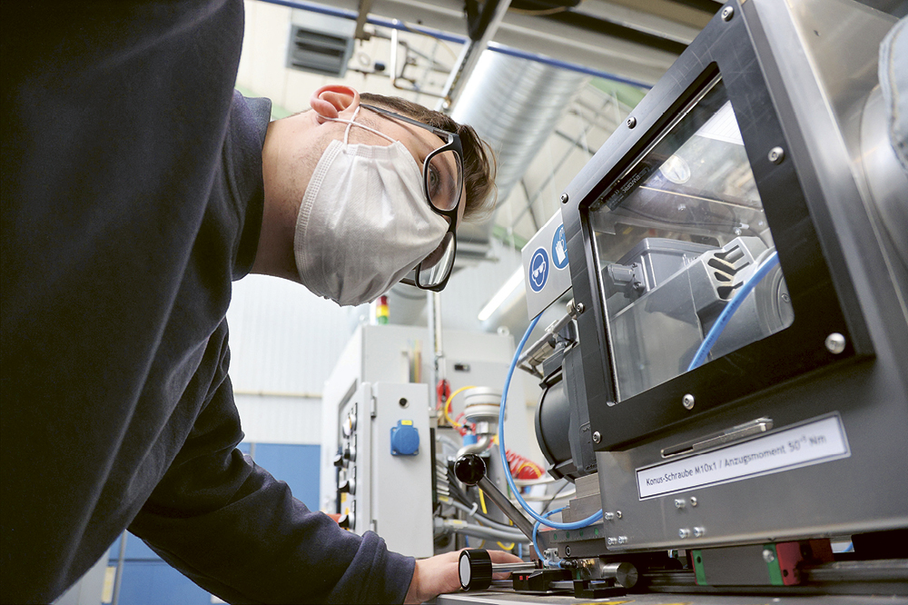 Ein Mann mit Maske schaut in einer Fabrik auf den Prüfstand einer Maschine.