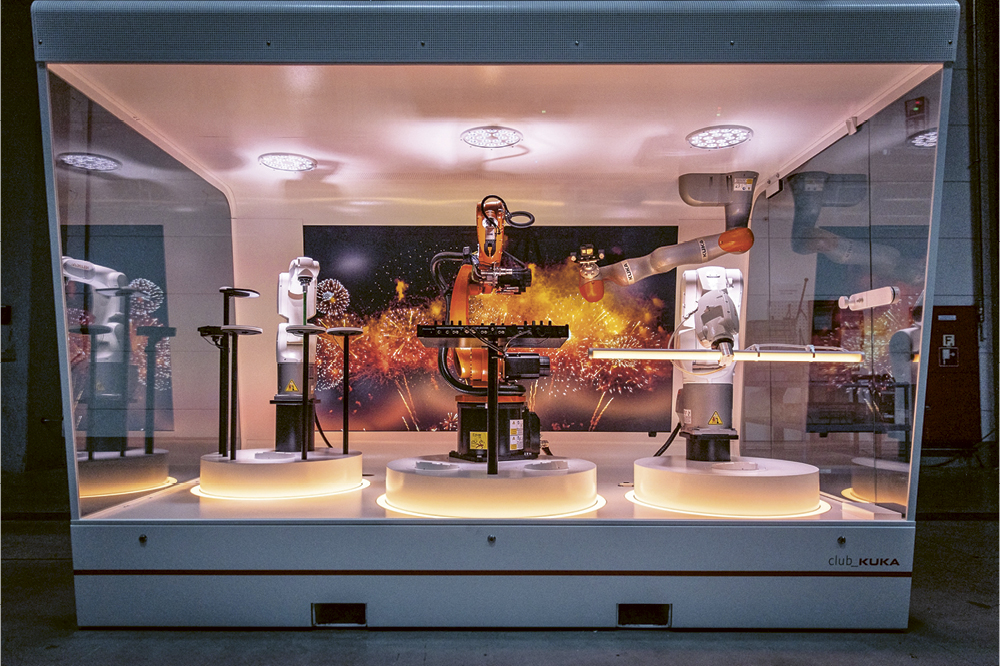 Roboter spielen in einem gläsernen Raum auf einer Showbhne Schlagzeug und Keyboard mit einem Bild von einem Feuerwerk im Hintergrund