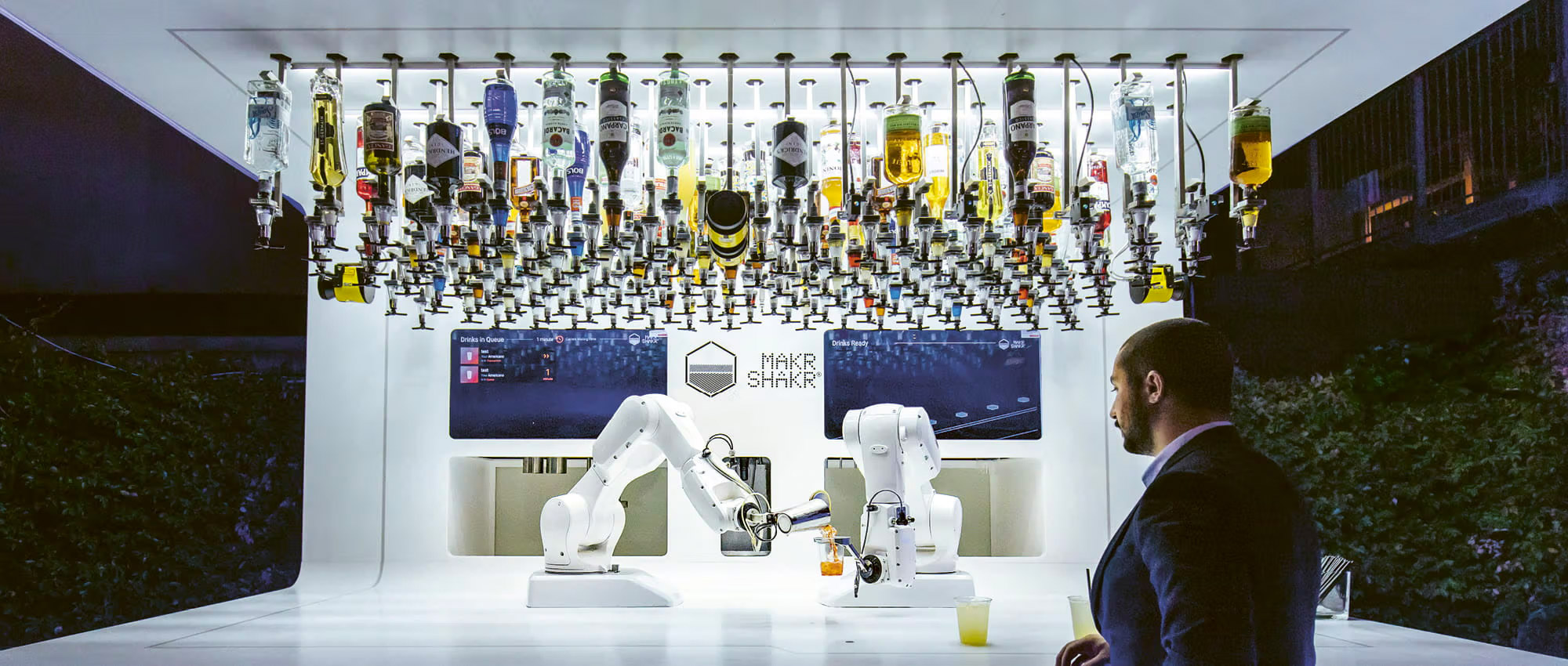 Ein Mann steht mit einem Cocktailbecher vor einem Kasten in dem zwei Roboterarme einen Cocktail mischen und mehreren verschiedenen Alkoholflaschen an der Decke. Copyright: KUKA Group
