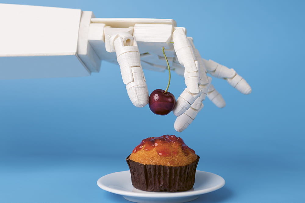 Eine weiße Roboterhand greift eine Kirsche und setzt sie auf einen Muffin. 
