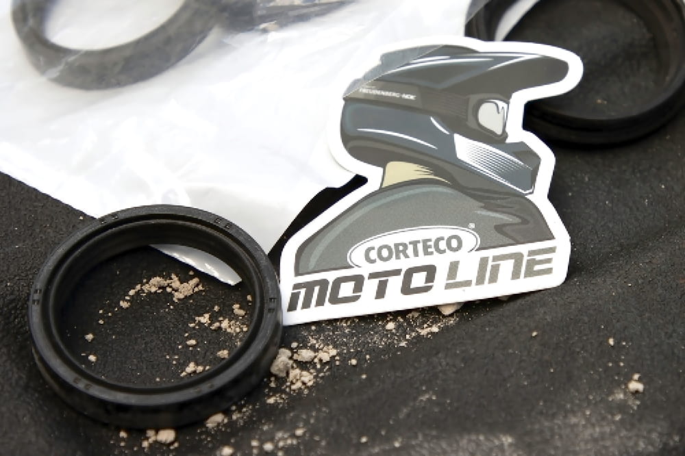 Ein Sticker von einer Person mit einem Motorradhelm auf dem Kopf mit der Aufschrift Corteco Moto Line liegt auf einem Tisch neben einer Dichtung. 