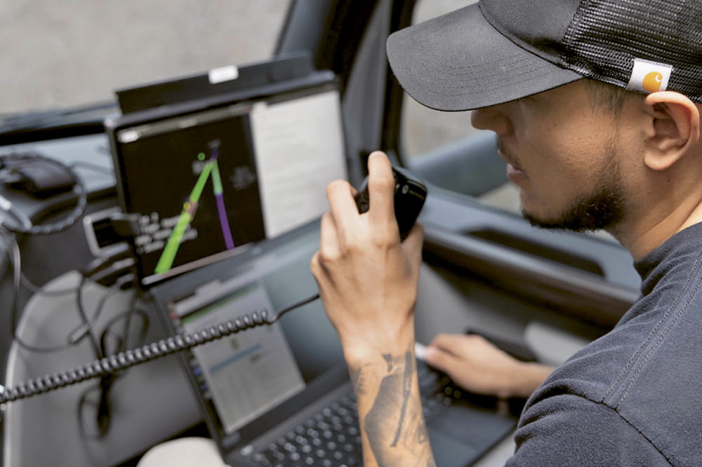Ein Mann sitzt in einem Auto an einem Computer und spricht in eine Gegensprechanlage.