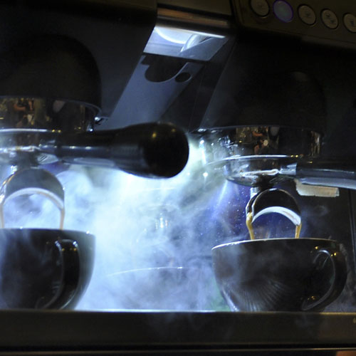 Nahaufnahme von zwei frisch gebrühten Espresso mit aufsteigendem Dampf aus einer Siebträgermaschine mit Beleuchtung im Hintergrund
