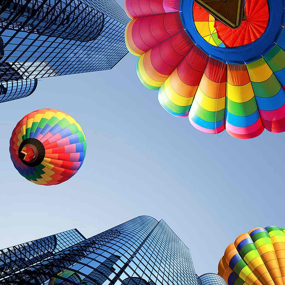 Drei zwischen gläsernen Hochhäusern aufsteigende, bunte Heißluftballons vor blauem Himmel 